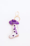 A small embroidered keychain featuring a white cowboy boot with purple floral embroidery and the word 'hardy' in purple script, alongside a purple flip-flop shoe, attached to a gold key ring on a white background.